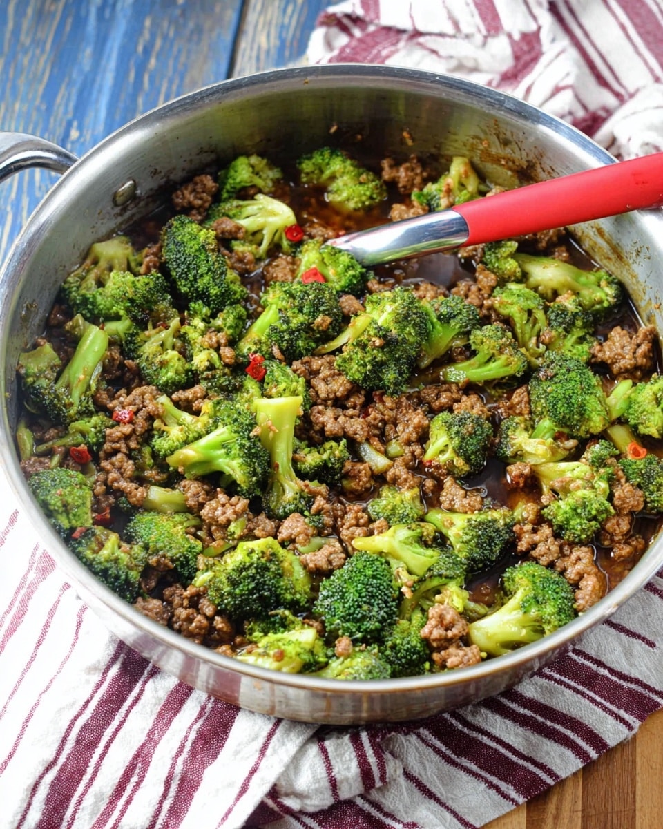 A silver cooking pan filled with a mix of small green broccoli florets and brown cooked ground meat in a shiny brown sauce, with some red specks scattered throughout. A red spoon with a silver handle rests inside the pan, positioned horizontally across the top. The pan is placed on a wooden surface covered partially by a cloth with white and dark red stripes. The background shows a blue wooden texture. Photo taken with an iphone --ar 4:5 --v 7