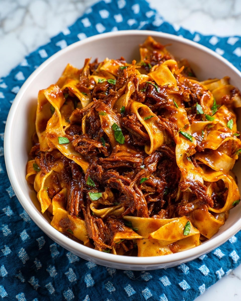 The image shows a white bowl filled with wide flat noodles that are coated in a thick, dark reddish-brown sauce. Mixed with the noodles are tender chunks of shredded meat that have a rich, saucy texture. Small pieces of green herbs are scattered throughout, adding pops of fresh color. The bowl is placed on a blue cloth with a diamond pattern, and the background is a white marbled texture. photo taken with an iphone --ar 4:5 --v 7