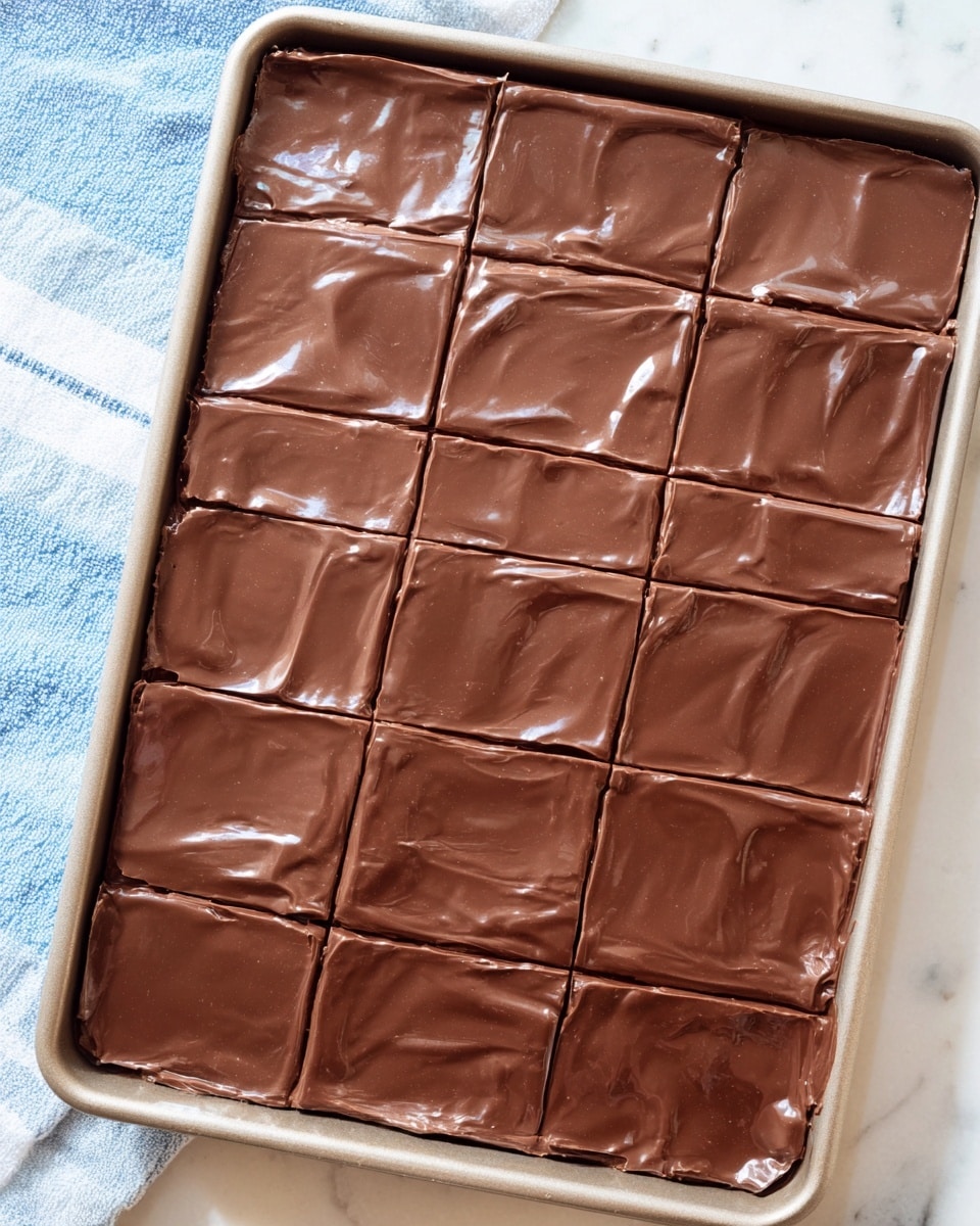 A rectangular tray filled with a smooth, shiny, dark brown chocolate layer covering the entire top. The chocolate has a slightly wavy texture with subtle reflections of light, making it look moist and glossy. The top layer is marked with even cuts, dividing the chocolate into 15 square pieces arranged in a 5 by 3 grid. The tray sits on a white marbled surface with a hint of a blue and white towel on the left side. Photo taken with an iphone --ar 4:5 --v 7