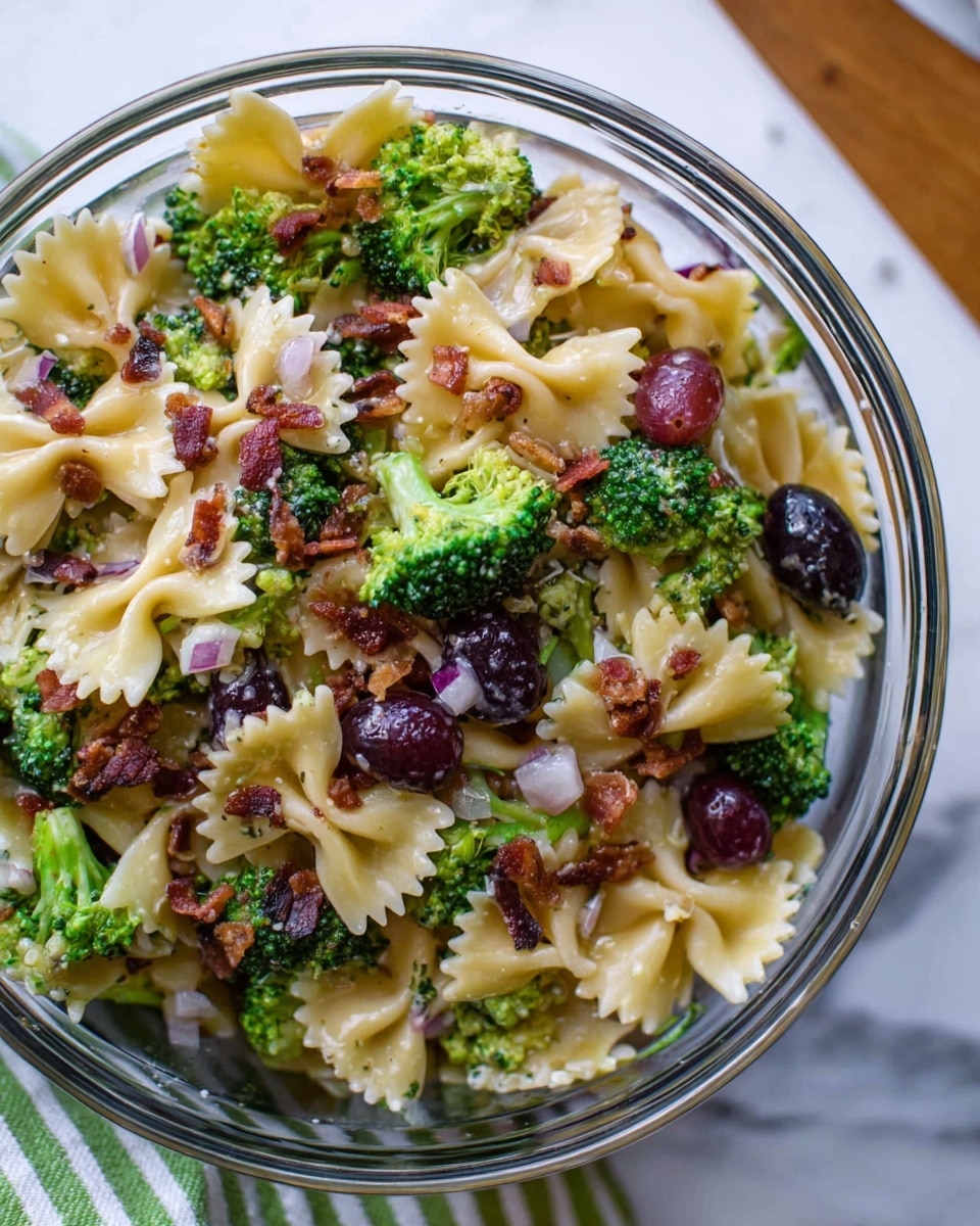 A clear glass bowl filled with a pasta salad made with many farfalle pasta pieces, mixed with green broccoli florets, small black grape halves, small white onion pieces, and crispy brown bacon bits scattered on top. The pasta is light yellow, the broccoli is bright green, the grapes are deep purple-black, and the onions are white and purple. The bowl is placed on a white marbled surface with a faint pattern of green and white stripes nearby. photo taken with an iphone --ar 4:5 --v 7