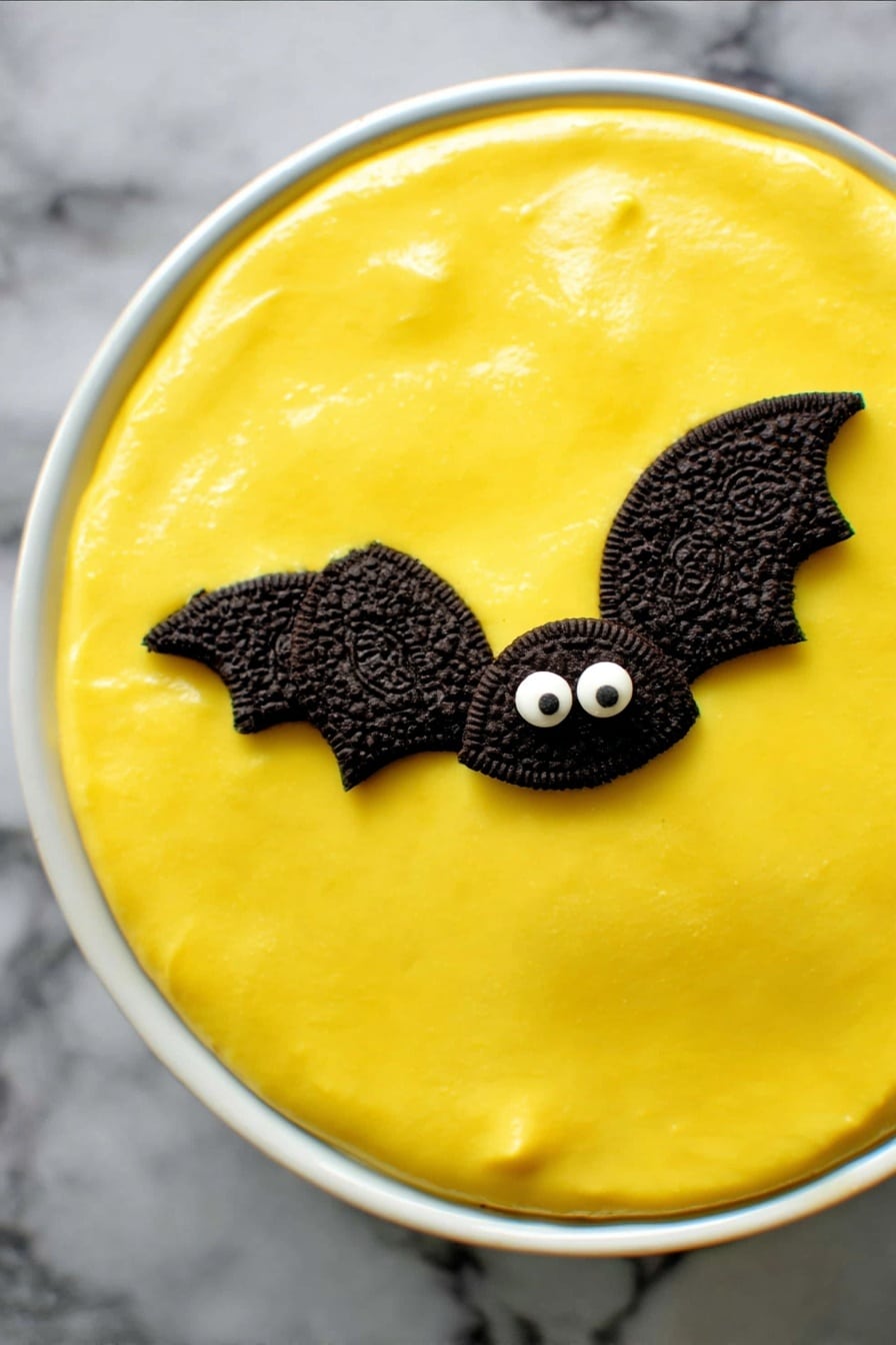 No Bake Boston Cream Halloween Pie, Halloween dessert recipes, festive no bake pies, easy Halloween desserts, spooky fall treat ideas - A round dessert in a white ceramic dish has a smooth, bright yellow top layer that looks creamy and shiny. On this yellow surface are two small decorations shaped like bats, made with halves of dark chocolate sandwich cookies for wings and two small white candy eyes on a chocolate base forming the face. The dish sits on a dark cloth background. photo taken with an iphone --ar 2:3 --v 7