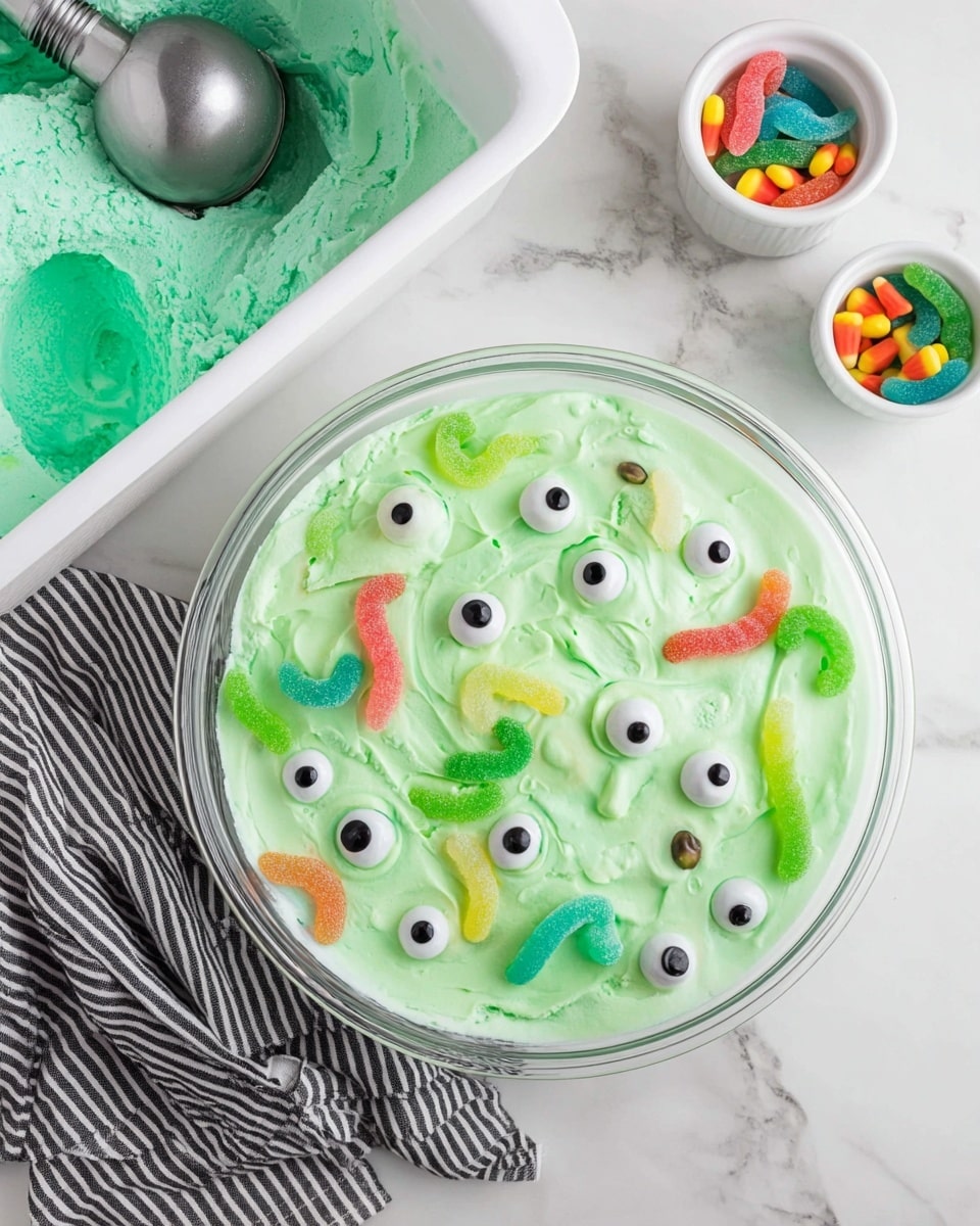 A clear glass bowl on a white marbled surface holds a bright light green, foamy mix with many pieces of candy eyeballs and gummy worms floating on top. Around the bowl, there is a white container filled with matching green ice cream that has a silver ice cream scoop with a black handle resting inside. Nearby, two small white bowls contain candy eyeballs and colorful gummy worms. The scene has a black and white striped cloth under the bowl for contrast. Photo taken with an iphone --ar 4:5 --v 7