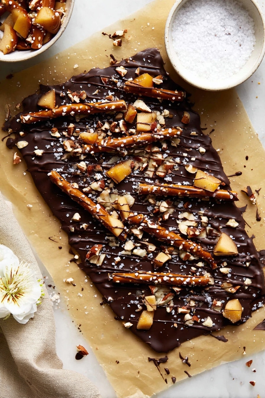 Caramel Apple Pretzel Bark, caramel apple pretzel bark, easy caramel bark, salty sweet pretzel bark, fall dessert recipes - The image shows thick dark chocolate bark with two main layers: a smooth, glossy dark brown chocolate base, and a thick topping layer of broken pretzel sticks, small light tan nut pieces, light tan apple chunks, and a drizzle of darker chocolate sauce across the top. The bark pieces rest on light brown parchment paper over a white marbled surface. To the side, there is a small white bowl filled with coarse sea salt. Small white flowers are placed decoratively around the edges. photo taken with an iphone --ar 2:3 --v 7