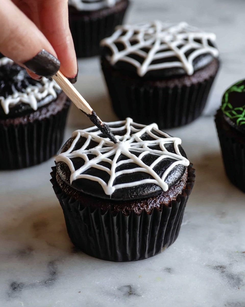 A dark chocolate cupcake in a black paper liner sits on a white marbled surface, topped with black frosting as the base layer. On top, white frosting forms a spider web pattern, with concentric circles connected by radial lines. A woman's hand holding a wooden toothpick is drawing details on the web, adding texture and depth. Other cupcakes with similar black and white spider web designs are blurred in the background. Photo taken with an iphone --ar 4:5 --v 7