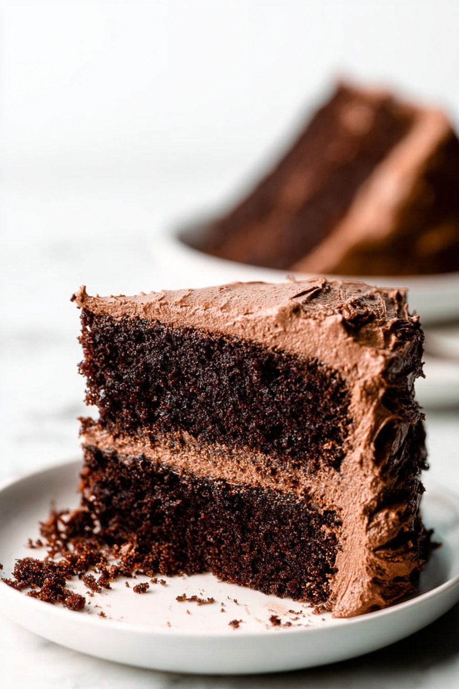 Gluten Free Vegan Chocolate Cake, dairy free vegan chocolate cake, eggless gluten free chocolate cake, vegan chocolate desserts, healthy chocolate cake recipe - A close-up view of a two-layer chocolate cake slice on a white plate, placed on a white marbled surface. The cake layers are dark brown and moist with a rough texture. Between the layers is a medium brown chocolate frosting that looks creamy and smooth. The outside of the cake slice is covered in the same chocolate frosting, which is thick and slightly swirled, with rough edges. Some frosting has spilled onto the plate. In the background, there is a blurred image of another chocolate cake slice on a white plate. Photo taken with an iphone --ar 2:3 --v 7