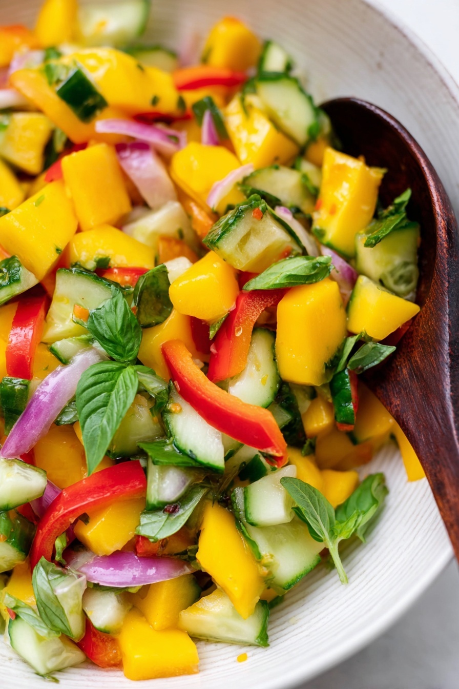 Mango Salad with Fresh Basil and Ginger Dressing, mango salad recipe, fresh fruit salad, healthy mango salad, tropical fruit salad - A close-up of a colorful salad in a white bowl with a white marbled background, showing bright yellow mango cubes, green cucumber pieces, red bell pepper chunks, thin slices of light purple onion, and fresh green basil leaves mixed together. A dark wooden spoon is resting inside the bowl, partly covered by the salad. The colors are vibrant and fresh, with the textures of the juicy mango, crunchy cucumber, and leafy greens clearly visible. Photo taken with an iphone --ar 2:3 --v 7