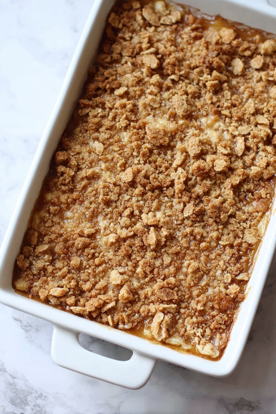 Pumpkin Dump Cake, easy fall dessert, pumpkin dessert recipe, quick dump cake, beginner friendly pumpkin treat - The image shows a baked dessert in a white rectangular dish with two handles. The dessert has one main layer with a golden brown crumbly topping made of crushed pieces with a rough, uneven texture. The color is mixed shades of light to medium brown, giving a crunchy and toasted look. The dish is placed on a white marbled surface. Photo taken with an iphone --ar 2:3 --v 7
