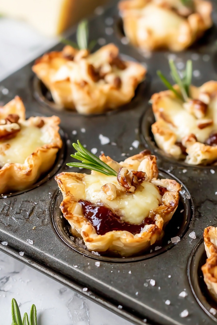 Cranberry Brie Bites without Puff Pastry, Cranberry Brie Bites, Holiday Appetizers with Cranberries and Brie, Easy Brie Bites for Parties, Puff Pastry Cranberry Brie Appetizer - The image shows mini tartlets baked in a dark metal muffin tray, each tartlet having three main layers. The bottom layer is a golden-brown, flaky pastry shaped into small cups with slightly crispy edges. The middle layer is a glossy, dark red jam or fruit preserve that peeks out from beneath the top. The top layer is a melted, creamy pale yellow cheese, topped with small brown nut pieces and a fresh green rosemary sprig for garnish. The background is a white marbled texture, and some salt granules are scattered around the tray. photo taken with an iphone --ar 2:3 --v 7