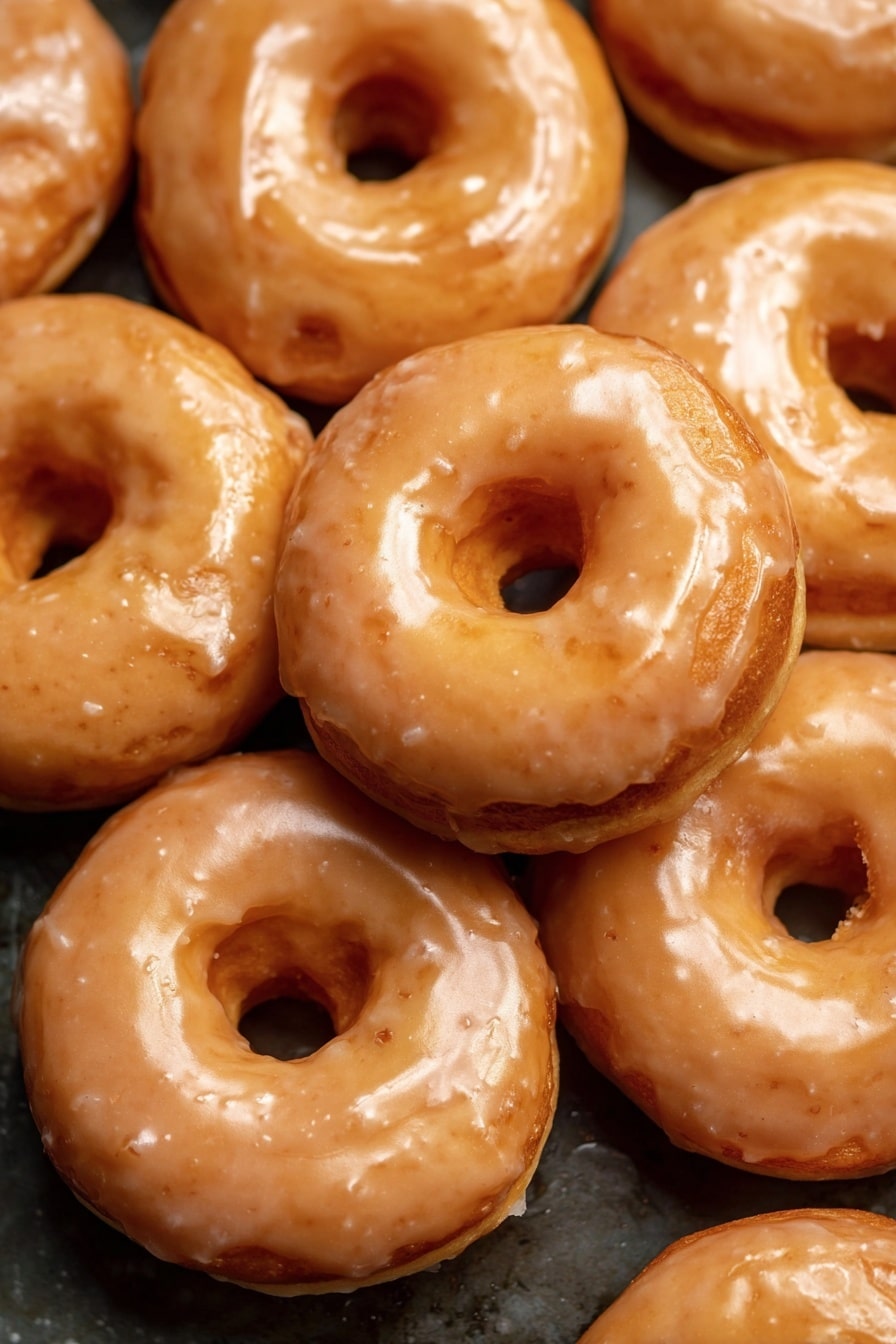 Vegan Pumpkin Donuts with Cinnamon Maple Glaze, vegan fall donuts, pumpkin spice vegan donuts, homemade vegan pumpkin donuts, plant-based pumpkin breakfast treats - The image shows a close-up of many round donuts stacked together. Each donut has two visible layers: a base layer that is a golden brown color with a soft texture and a shiny glaze layer on top that is light tan and slightly translucent. The glaze evenly covers the top and sides of each donut, giving them a glossy look. The donuts have a round hole in the middle, and their surface shows slight bumps and smooth spots. The background is a dark surface contrasting with the shiny donuts. photo taken with an iphone --ar 2:3 --v 7