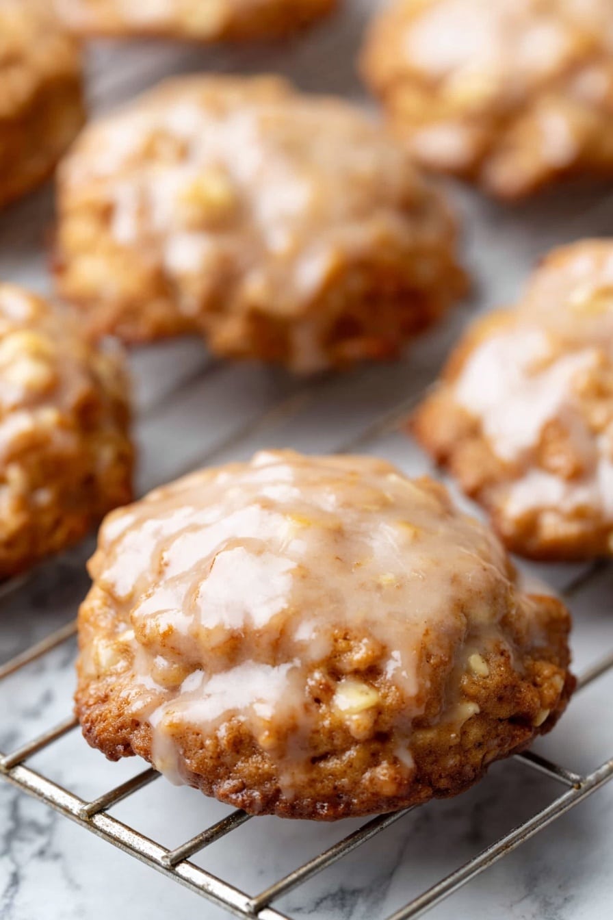 Crispy Apple Fritters with Vanilla Glaze, apple fritter recipe, fall dessert ideas, homemade apple fritters, easy fritter recipe - The image shows several round cookies on a cooling rack. Each cookie has a shiny, light brown glaze on top with a slightly uneven texture. The cookies have a golden brown color, with a rough surface and visible small pieces inside, likely nuts or oats. The cooling rack underneath is metal and placed over a white marbled surface. The cookies look soft and have a slightly wavy edge. Photo taken with an iphone --ar 2:3 --v 7