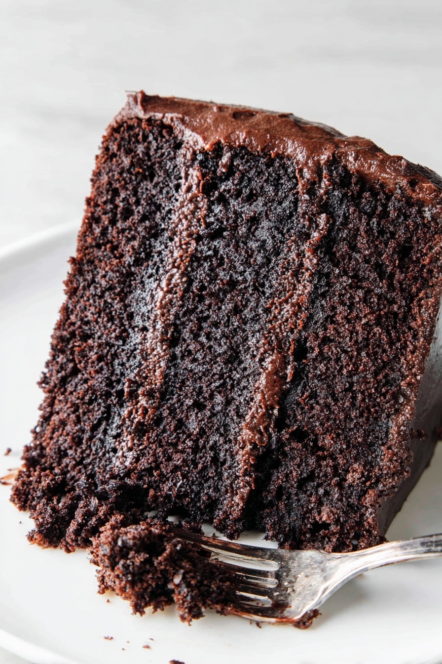 The Most Amazing Chocolate Cake, best chocolate cake recipe, moist chocolate cake, rich chocolate cake, homemade chocolate cake - The image shows a slice of chocolate cake on a white plate placed on a white marbled surface. The cake has two thick layers of moist, dark brown chocolate sponge with a rich, glossy chocolate frosting layer in the middle. The outside of the cake is covered with a smooth, shiny chocolate frosting. A silver fork with some chocolate frosting rests on the plate next to the slice. Photo taken with an iphone --ar 2:3 --v 7
