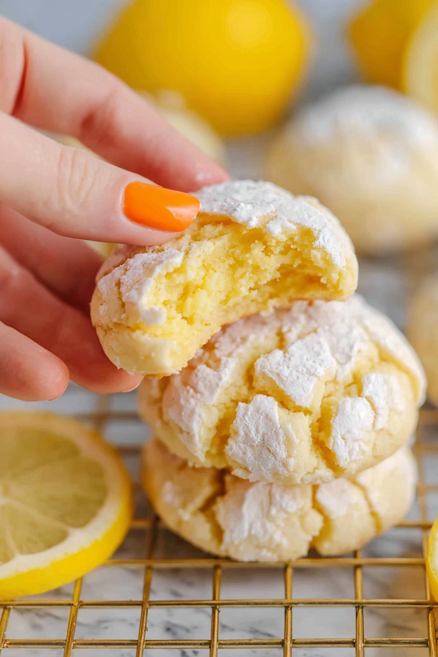 Lemon Crinkle Cookies, Lemon Crinkle Cookies Recipe, Bright Lemon Cookies, Soft Lemon Cookies, Zesty Lemon Cookies - A close-up image shows a woman's hand with orange nail polish holding a lemon cookie with a bite taken out of it, revealing its soft yellow inside. The cookie has a cracked top covered lightly with white powdered sugar. Underneath are more similar cookies stacked on a gold cooling rack. Around the rack are bright yellow lemon halves placed on a white marbled surface, adding fresh color to the scene. The photo taken with an iphone --ar 2:3 --v 7