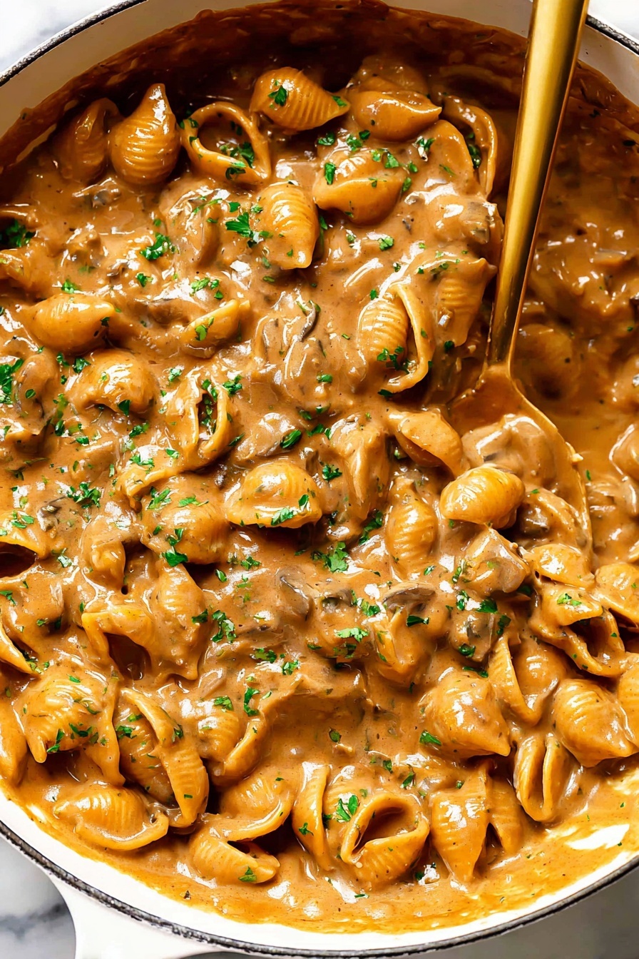 French Onion Pasta, caramelized onion pasta, easy French onion pasta recipe, comforting pasta dishes, savory onion pasta - A close-up view of a white pan filled with a rich, creamy mushroom sauce that has a warm brown color and a thick texture. The sauce contains many small mushroom slices that create a bumpy surface, covered evenly in the glossy, smooth sauce. Bright green parsley bits are scattered on top, adding a touch of color contrast. A shiny golden spoon rests inside the pan, partially submerged in the sauce, showing the thick consistency as it drags some sauce along. The background features a white marbled surface. photo taken with an iphone --ar 2:3 --v 7