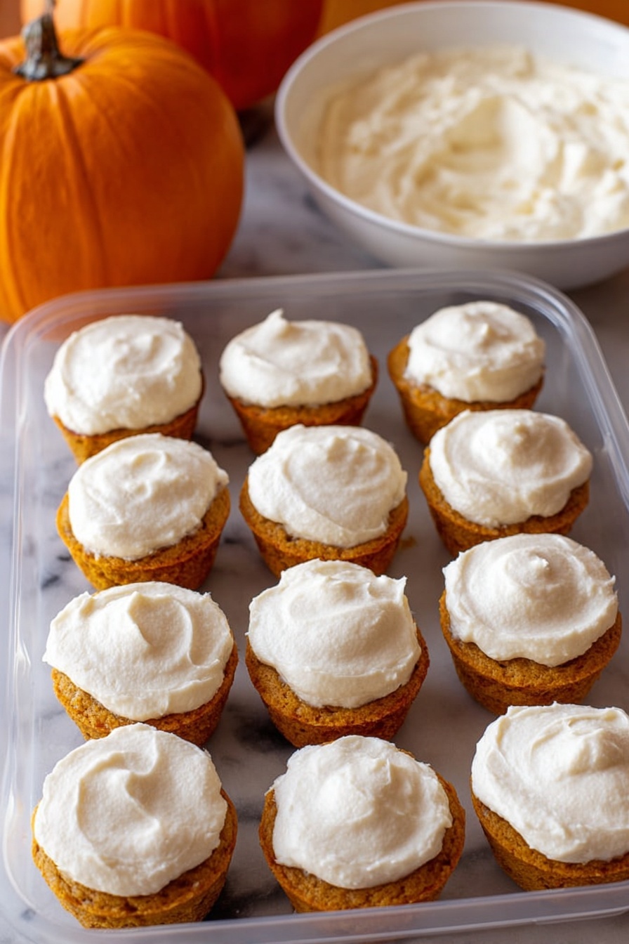 Flourless Pumpkin Muffins, pumpkin muffins without flour, healthy pumpkin muffin recipe, quick pumpkin muffin ideas, moist pumpkin muffin recipe - The image shows a clear plastic container filled with twelve small cupcakes arranged in three rows of four. Each cupcake has two layers: a golden orange base with a slightly crumbly texture and a thick layer of white frosting spread smoothly on top. The frosting covers the entire top surface of each cupcake with a slightly swirled pattern. In the background, there is an orange pumpkin on the left and a white bowl filled with more white frosting on the right, all placed on a white marbled surface. photo taken with an iphone --ar 2:3 --v 7