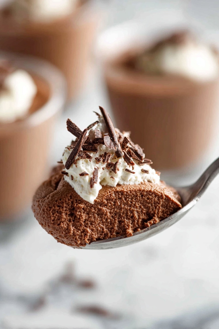 Decadent Chocolate Mousse, rich chocolate mousse recipe, silky chocolate mousse, easy chocolate mousse, homemade chocolate mousse - The image shows a clear glass filled with a rich, smooth dark brown chocolate pudding. On top, there is a dollop of white whipped cream with delicate dark chocolate shavings scattered over it. The glass is placed on a white marbled surface, and in the background, there are blurred similar glasses filled with the same dessert. Photo taken with an iphone --ar 2:3 --v 7