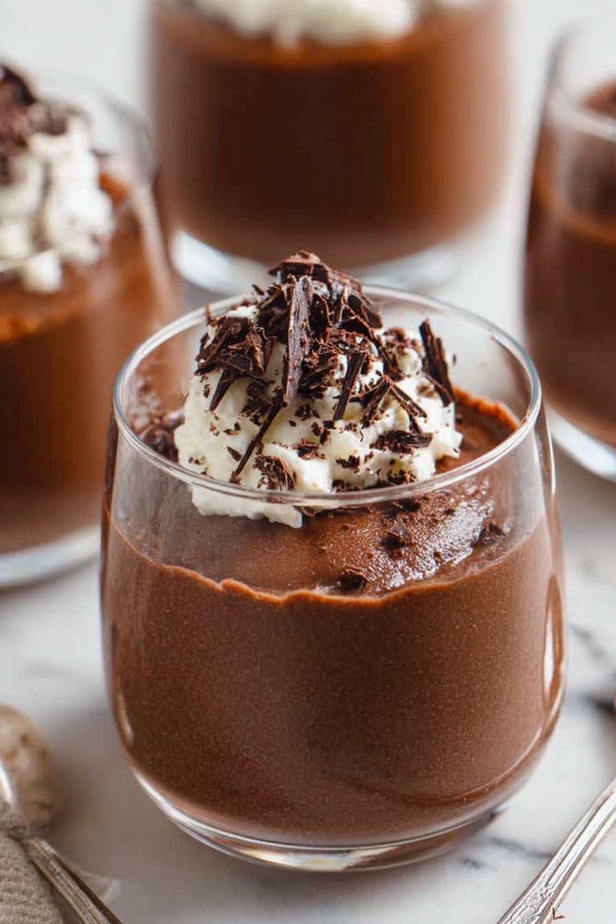 Decadent Chocolate Mousse, rich chocolate mousse recipe, silky chocolate mousse, easy chocolate mousse, homemade chocolate mousse - A close-up of a spoon holding a scoop of light and airy chocolate mousse with a rough texture, topped with a small dollop of white whipped cream and thin, dark chocolate shavings scattered on top, with two blurry white bowls filled with the same mousse and cream in the background, all set on a white marbled surface. photo taken with an iphone --ar 2:3 --v 7