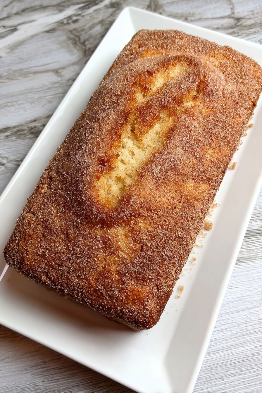 Cinnamon Donut Bread, cinnamon donut bread, cinnamon bread recipe, sweet breakfast bread, cinnamon swirl loaf - A rectangular cake with a golden-brown top layer covered evenly with a fine, grainy light brown sugar coating sits on a white rectangular plate. The cake's top surface shows a gently cracked texture with soft ridges and swirls, revealing a tender and moist interior below the sugar crust. The plate rests on a white marbled texture surface. photo taken with an iphone --ar 2:3 --v 7