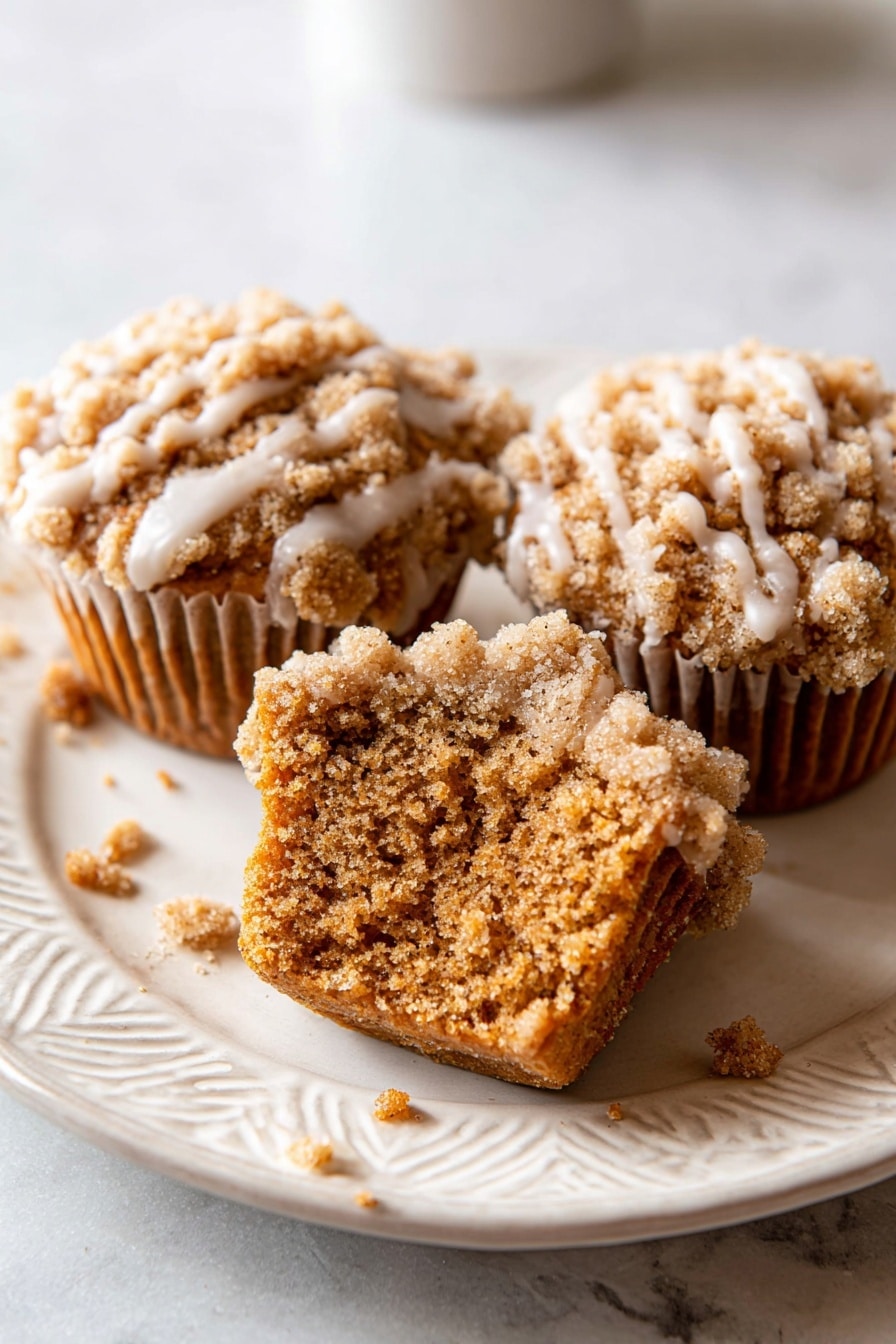 Pumpkin Crumb Cake Muffins, fall muffin recipes, pumpkin spice muffins, easy pumpkin muffins, moist pumpkin breakfast muffins - The image shows seven golden brown muffins arranged closely on a white plate. Each muffin has a crumbly, rough-textured top layer that is light brown and covered with a light cream-colored glaze that drips down the sides in thin streaks. The muffins are placed on a smooth white marbled surface underneath the plate. Some crumbs are scattered around the plate and surface. The lighting highlights the texture and glaze shine on the muffins. Photo taken with an iphone --ar 2:3 --v 7