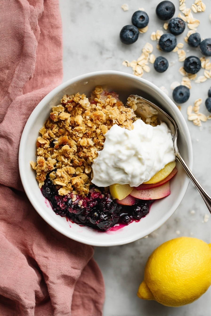 Apple and Blueberry Crumble, fruit crumble recipe, easy berry dessert, baked fruit crisp, cozy fall desserts - A white bowl sits on a white marbled surface, filled with three main layers. The bottom layer contains dark blue and purple berries mixed with thin slices of light pink fruit. Above that is a thick, crumbly layer of golden brown oat crumble with hints of toasted oats on top. The final layer is a generous dollop of white whipped cream placed in the center of the crumble. A silver spoon is partly inside the bowl on the right side. Next to the bowl is a yellow lemon and scattered blueberries and oats, with a soft pink cloth to the left. photo taken with an iphone --ar 2:3 --v 7