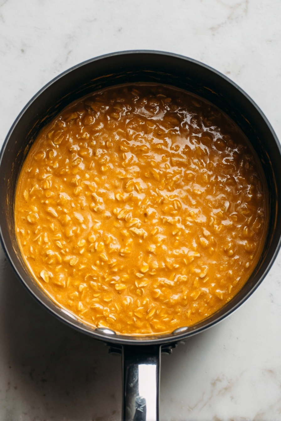 Pumpkin Oatmeal, pumpkin oatmeal recipe, cozy breakfast, autumn breakfast ideas, healthy pumpkin oatmeal - This image shows a close-up top view of a round black saucepan filled with a thick, creamy orange mixture with visible small oats throughout. The oats give the surface a bumpy texture, and the mixture looks smooth and warm. The saucepan is placed on a white marbled surface, and the shiny black handle is visible at the bottom right of the image. The lighting highlights the glossy shine of the mixture, making it look fresh and appetizing. photo taken with an iphone --ar 2:3 --v 7