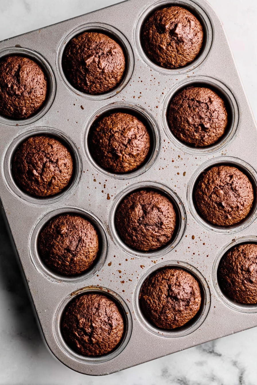 Sticky Toffee Pudding with Warm Toffee Sauce, British dessert recipes, moist date pudding, classic toffee dessert, indulgent dessert recipes - A metal muffin tray with 12 evenly spaced round muffins baked inside, each muffin a deep brown color with a rough, slightly cracked top texture showing some small chunks within. The muffins fit neatly in each circular slot of the tray, which shows small spots of light brown residue from baking. The tray is placed on a white marbled surface with soft natural lighting highlighting the muffins' golden and dark brown hues. photo taken with an iphone --ar 2:3 --v 7