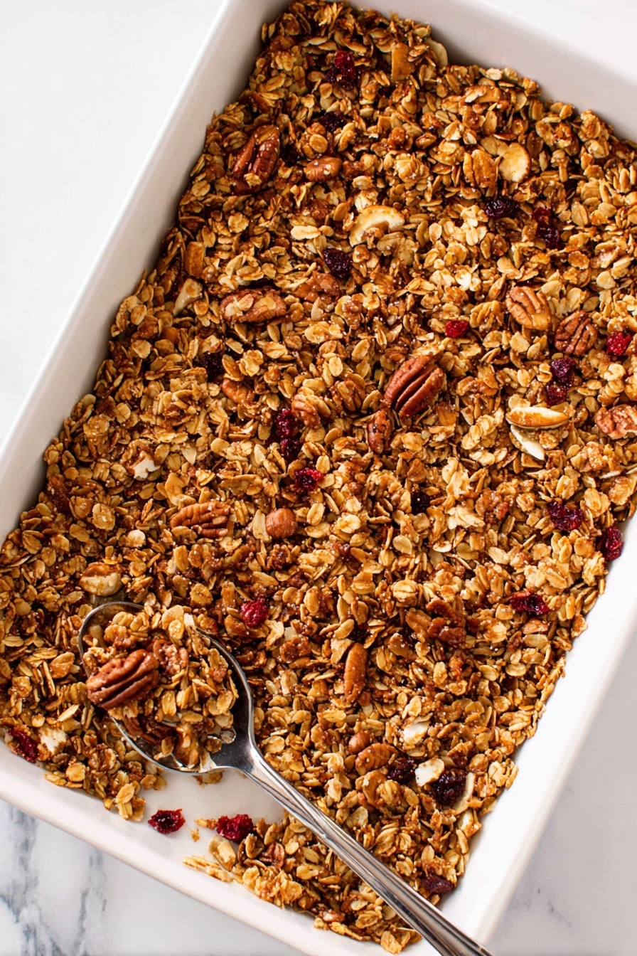 Gingerbread Granola, spiced granola recipe, crunchy breakfast granola, homemade gingerbread granola, holiday granola snack - A deep white rectangular baking tray filled with a golden brown granola mix that includes oats, nuts like pecans, and small red dried fruit pieces scattered evenly throughout. The granola layer looks crunchy with a mix of small and larger clusters, and a silver spoon rests on the bottom left corner inside the tray, holding some granola pieces. The background is a white marbled surface. Photo taken with an iphone --ar 2:3 --v 7
