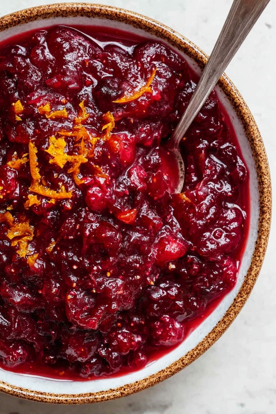 Easy Cranberry Sauce with Orange Juice, cranberry sauce, orange cranberry appetizer, holiday cranberry sauce, homemade fruit sauce - A close-up view of a thick, deep red cranberry sauce with a chunky texture, showing visible pieces of fruit mixed in. Small bits of bright orange zest are scattered on top, adding color contrast. The sauce fills a white bowl with brown speckled edges, and a silver spoon is placed inside the bowl, partly submerged in the sauce. The bowl sits on a white marbled surface. photo taken with an iphone --ar 2:3 --v 7