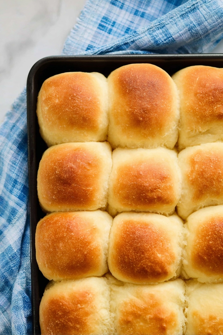 Vegan Dinner Rolls, Fluffy Vegan Roll Recipe, Plant-Based Dinner Rolls, Soft Vegan Bread Rolls, Easy Vegan Dinner Rolls - The image shows a close-up top view of sixteen golden-brown, fluffy bread rolls arranged in a 4 by 4 grid inside a black baking tray. Each roll has a slightly rounded top with a soft, smooth texture and a light crust that is golden on the upper part, fading to creamy white around the sides. The baking tray is placed on a blue and white checkered cloth on a white marbled surface. The rolls touch each other, showing their soft and puffy sides. photo taken with an iphone --ar 2:3 --v 7