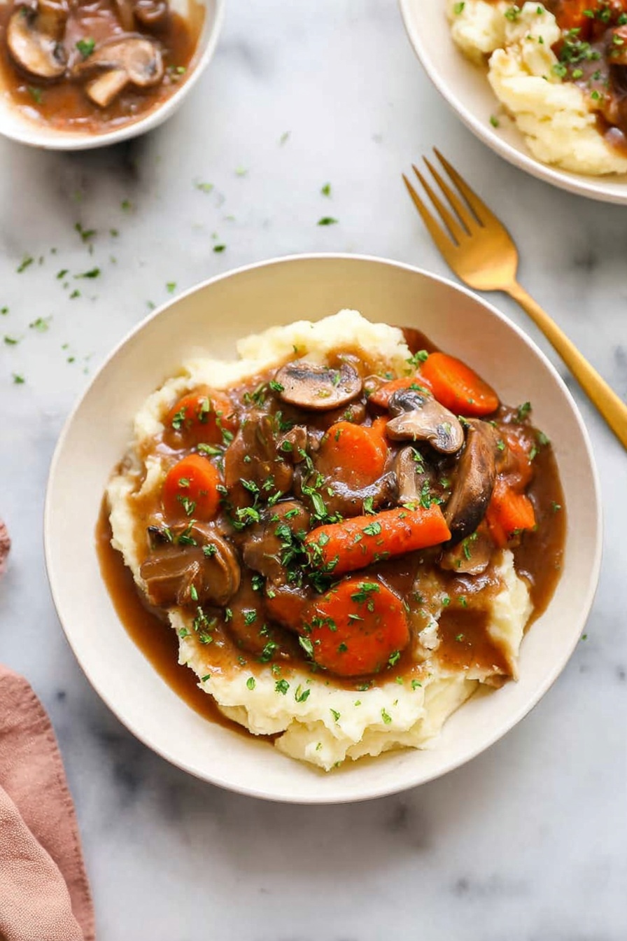 Mushroom Bourguignon, vegan mushroom stew, plant-based French stew, hearty mushroom recipes, vegetarian bourguignon - A white bowl holds a dish with two main layers: the bottom layer is creamy mashed potatoes, soft and pale in color, covering the base of the bowl. On top is a rich brown gravy mixed with sliced mushrooms and large carrot chunks, with chopped green herbs sprinkled all over for color. A golden spoon and fork sit on the white marbled surface beside the bowl. In the background, another white bowl with the same dish is barely visible. The lighting is bright and natural, highlighting the textures and colors of the food photo taken with an iphone --ar 2:3 --v 7