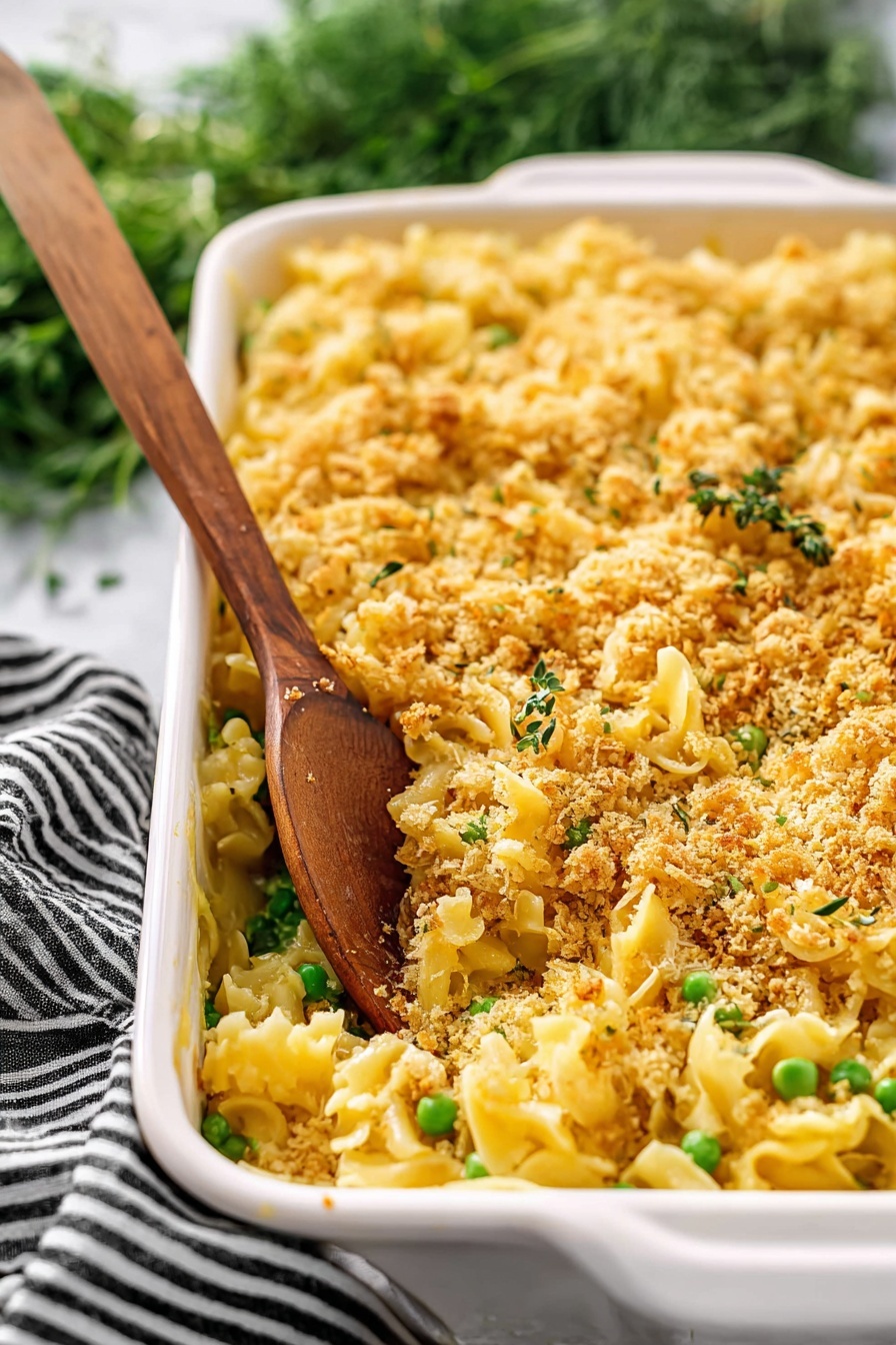 Creamy Chicken Casserole, Chicken casserole recipe, cheesy chicken bake, easy chicken dinner, comforting casserole dishes - The image shows a white rectangular baking dish filled with a pasta casserole. The dish has three main layers: a bottom layer of yellow pasta, a middle layer with green peas mixed throughout, and a top layer covered with a golden brown crumbly topping that looks crispy. A wooden spoon stands inside the dish, scooping some pasta and crumbs. The background features a white marbled surface and some green herbs with a black and white striped cloth nearby. photo taken with an iphone --ar 2:3 --v 7