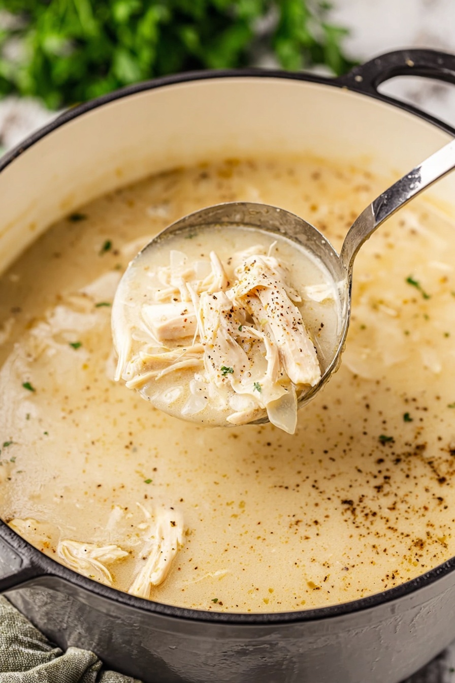 Southern Style Chicken and Dumplings, Southern Chicken and Dumplings, Classic Southern Comfort Food, Easy Chicken and Dumplings Recipe, Hearty Chicken and Dumplings - A close-up view of a creamy light beige soup with shredded light tan chicken pieces mixed in. The soup has a smooth, thick texture with visible specks of black pepper spread on the surface. A shiny metal ladle lifts a portion of the soup showing the creamy broth and chicken clearly. The soup pot is white inside with a black exterior handle visible, resting on a white marbled surface with green blurred herbs in the background. Photo taken with an iphone --ar 2:3 --v 7