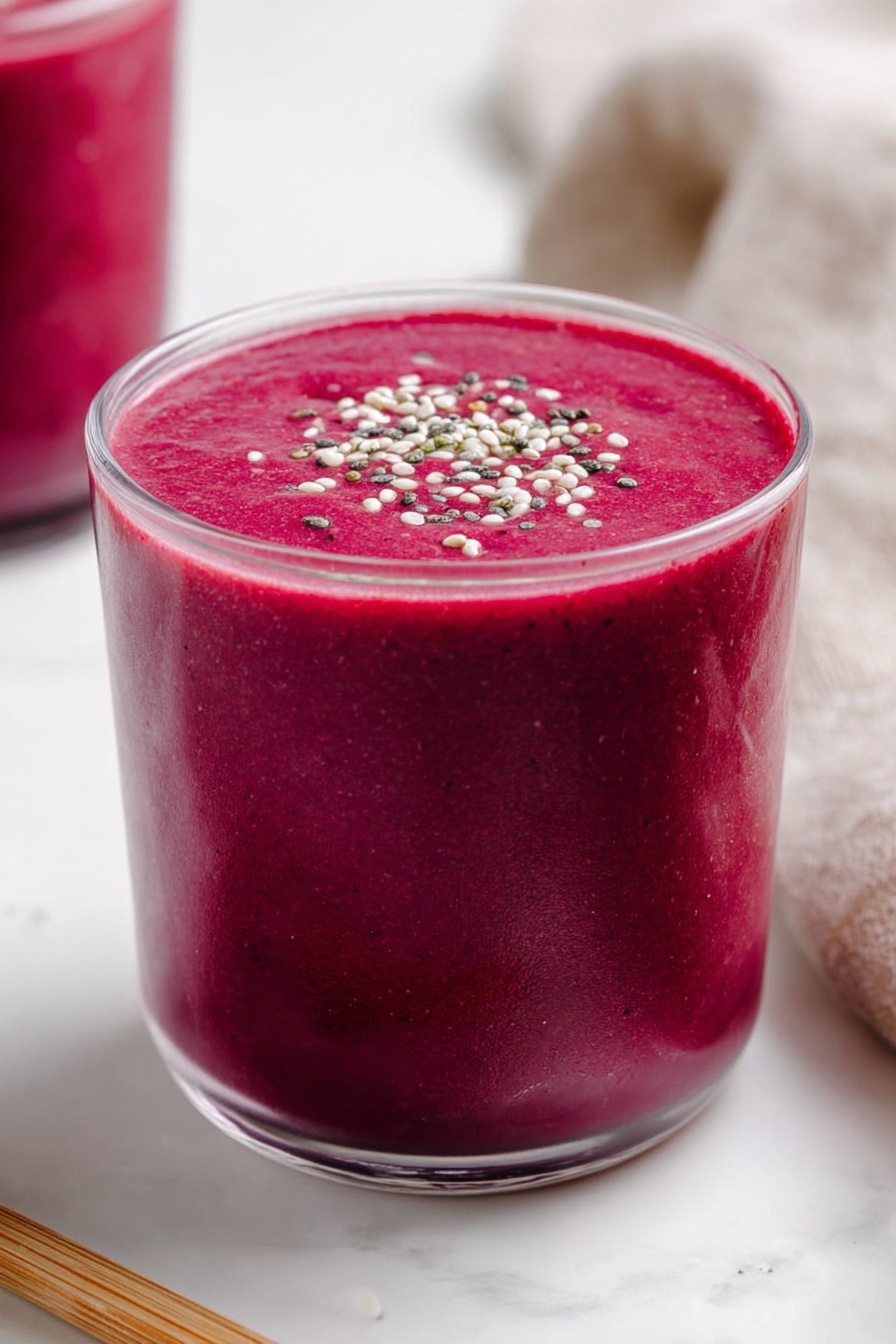 Banana Beet Smoothie, healthy beet smoothie, nutritious smoothie recipes, fruit and vegetable smoothie, easy breakfast smoothies - A clear glass is filled with a thick, vibrant dark pink smoothie, almost reaching the top with a smooth and creamy texture. The smoothie surface is flat and topped with small white, black, and green seeds or grains sprinkled evenly across the center. The glass rests on a white marbled surface, with part of another glass filled with the same pink smoothie visible blurred in the background on the left. A light brown wooden stick lies horizontally near the base of the main glass on the left side, and a soft beige cloth is partly visible on the right side. Photo taken with an iphone --ar 2:3 --v 7