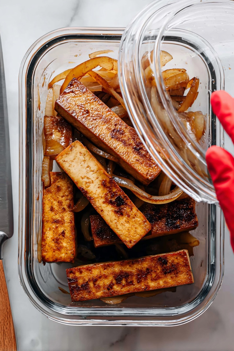 Sautéed Lime Tofu, lime tofu stir-fry, quick vegetarian dinner, tangy tofu recipe, plant-based tofu dishes - The image shows a clear glass container holding several thick rectangular pieces of cooked tofu that are brown and have a slightly charred texture on some edges. Mixed among the tofu pieces are translucent sautéed onion slices that are light brown and curled. The container is placed on a white marbled surface, and a woman's hand with a red-gloved fingertip is lifting the clear lid. To the left, a silver knife rests on the surface partially visible. photo taken with an iphone --ar 2:3 --v 7