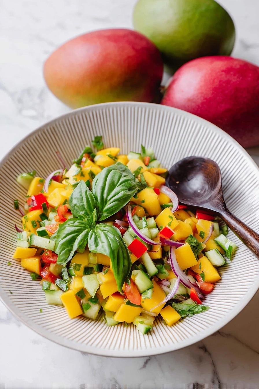 Mango Salad with Fresh Basil and Ginger Dressing, mango salad recipe, fresh fruit salad, healthy mango salad, tropical fruit salad - A white bowl with fine vertical ridges holds a colorful mango salad. The salad has many small diced yellow mango pieces mixed with green cucumber, red bell pepper chunks, and thin slices of purple onion. There are several fresh green basil leaves on top and mixed throughout the salad. A dark wooden spoon rests inside the bowl on the right side. The bowl is set on a white marbled surface with two whole mangoes, one red and one green, placed slightly blurred in the background. Photo taken with an iphone --ar 2:3 --v 7