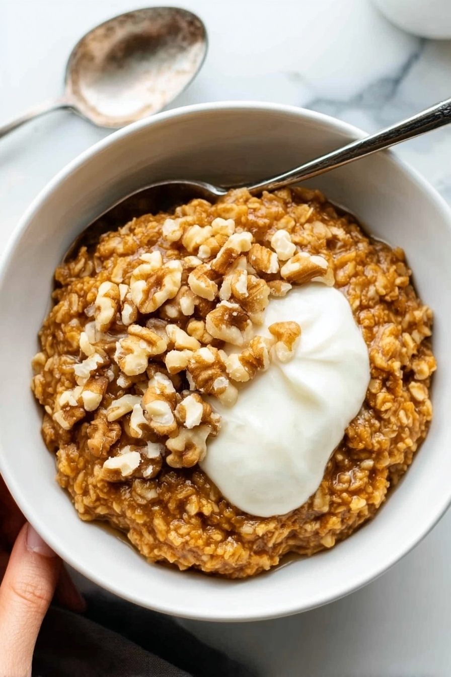 Pumpkin Oatmeal, pumpkin oatmeal recipe, cozy breakfast, autumn breakfast ideas, healthy pumpkin oatmeal - A white bowl filled with thick oatmeal that is golden brown in color and has a textured look from large oats. On top of the oatmeal, there is a layer of whole walnut pieces, adding a rough, uneven surface in light brown shades. Next to the walnuts, there is a dollop of smooth, white cream sitting on the right side of the bowl. The bowl is set on a white marbled surface, and a silver spoon rests next to it on the left. In the upper left corner, a woman's hand is visible holding the bowl. Photo taken with an iphone --ar 2:3 --v 7