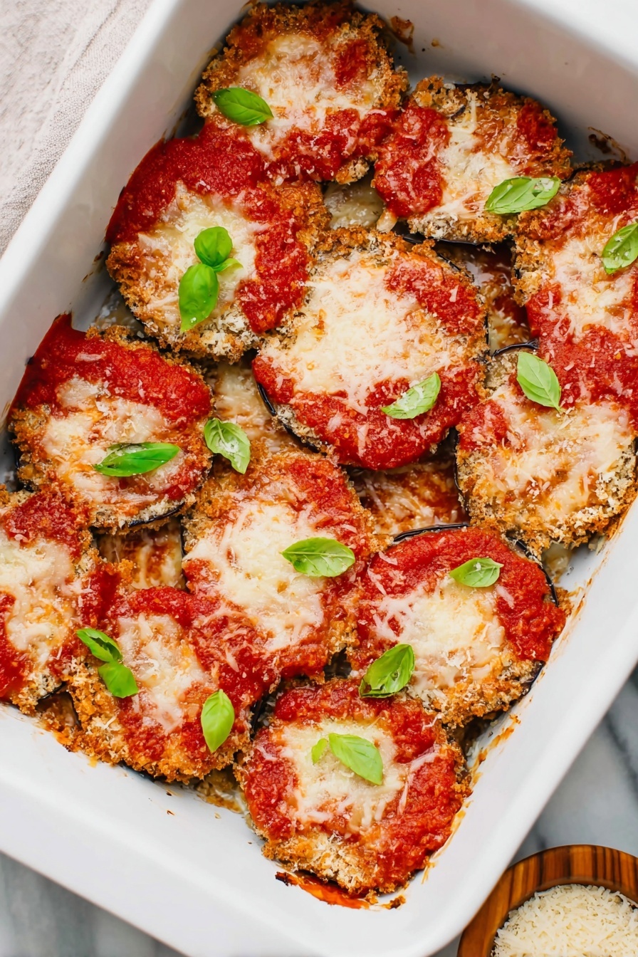 Vegan Eggplant Parmesan, plant-based Eggplant Parmesan, dairy-free Eggplant Parmesan, cheesy vegan Eggplant Parmesan, crispy eggplant dairy-free - The image shows a white baking dish filled with one layer of round slices of breaded eggplant, each topped with a bright red tomato sauce patch and a generous layer of melted light golden cheese. Fresh small green basil leaves are scattered on top, adding a touch of vibrant color. The eggplant slices have a crispy textured brown crust visible at the edges. A small wooden bowl with grated cheese is placed nearby on a white marbled surface. Photo taken with an iphone --ar 2:3 --v 7