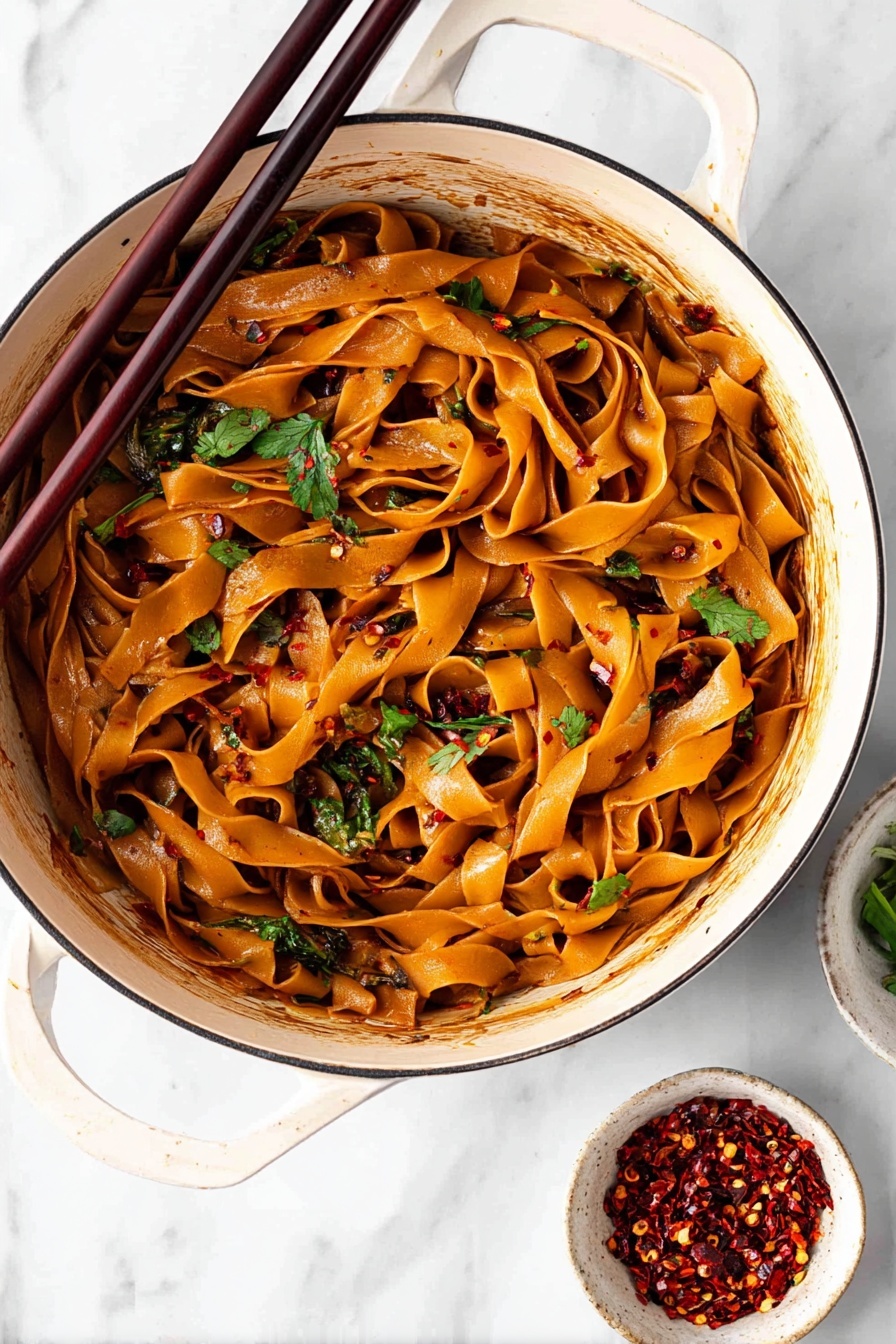 Spicy Chili Garlic Noodles, chili garlic noodle dish, spicy noodle recipe, quick Asian noodles, flavorful chili garlic stir-fry - A white bowl filled with wide flat noodles coated in a shiny reddish-brown sauce is shown from above. The noodles are mixed and curled inside the bowl with visible pieces of fresh green onions and chopped cilantro scattered on top. There are small white sesame seeds and dark chili flakes sprinkled over the noodles. A pair of dark wooden chopsticks rests on the edge of the bowl leaning inward. In the blurred background, two small white bowls hold red chili flakes and more chopped green onions. The surface is a white marbled texture. photo taken with an iphone --ar 2:3 --v 7