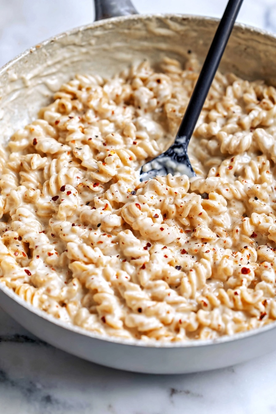 Creamy Cottage Cheese Pasta Sauce, healthy pasta sauce, light creamy pasta, high protein pasta toppings, quick cottage cheese pasta - The image shows a large gray bowl filled with a creamy, light beige pasta dish. The pasta is rotini, with a spiral shape, fully coated with a thick, smooth sauce that has a cheesy and slightly speckled texture. A woman's hand holding a metal spoon is stirring the pasta, lifting a spoonful from the center. The bowl sits on a surface with a white marbled texture. The overall look is warm and creamy, with the pasta fully covered in the sauce to give it a rich appearance. photo taken with an iphone --ar 2:3 --v 7