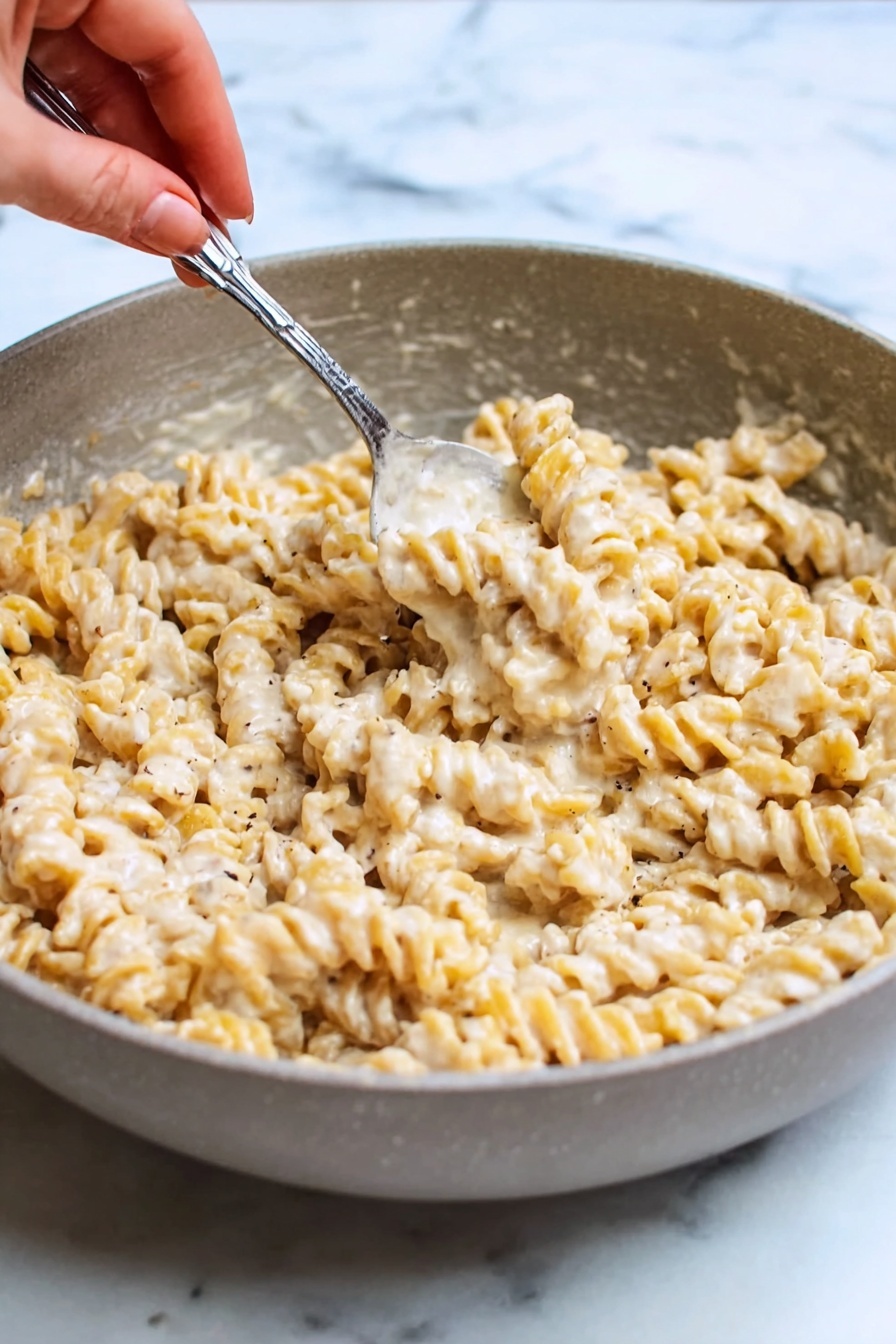Creamy Cottage Cheese Pasta Sauce, healthy pasta sauce, light creamy pasta, high protein pasta toppings, quick cottage cheese pasta - A close-up view of a white pan filled with creamy spiral pasta coated in a thick, smooth, off-white sauce with visible small black pepper and red chili flakes mixed in. A black spoon is standing upright in the middle of the dish, slightly submerged in the pasta. The pan sits on a white marbled surface. The overall look is warm and rich with a soft texture on the pasta photo taken with an iphone --ar 2:3 --v 7
