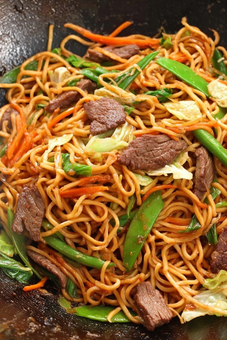 Beef Lo Mein Stir Fry, quick beef stir fry recipes, easy Asian noodles, flavorful beef stir fry, weeknight dinner ideas - This image shows a close-up of a wok filled with stir-fried noodles, meat slices, and mixed vegetables. The noodles are long and orange-brown, forming the main layer twisted together. Scattered throughout are medium-brown pieces of cooked meat, adding a rich texture. Bright green vegetables like sliced green onion and snap peas are placed evenly, with thin strips of orange carrot and light green cabbage mixed in, adding new colors and crunch. The surface inside the wok is dark and slightly shiny, highlighting the fresh, vibrant stir-fry. photo taken with an iphone --ar 2:3 --v 7