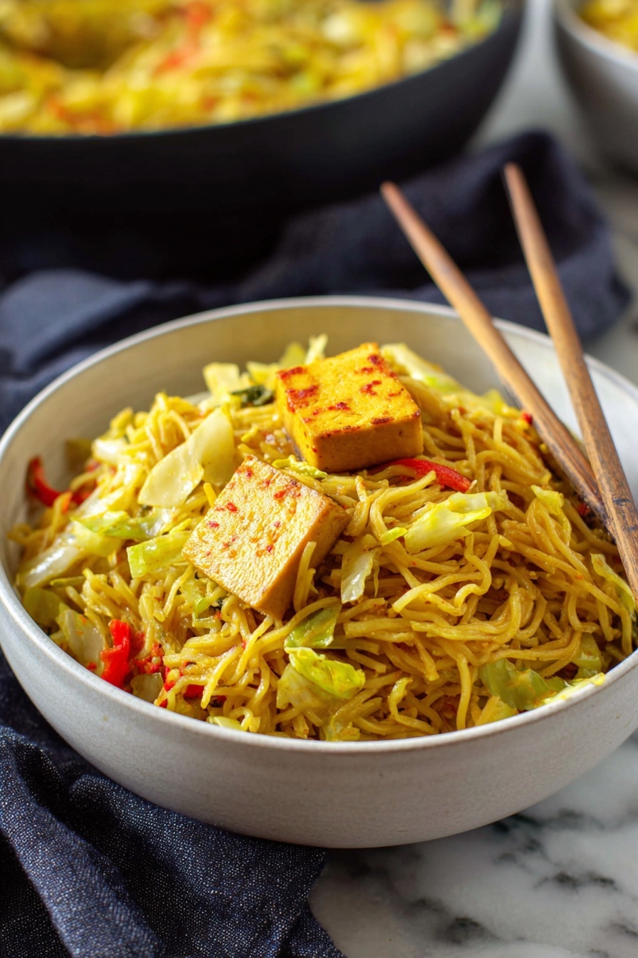 Quick and Easy Singapore Noodles, Singapore Noodles, Stir-Fry Noodles, Asian Noodle Recipes, Fast Weeknight Dinner - A deep white bowl full of thin yellow noodles mixed with pieces of green cabbage and small bits of red bell pepper, topped with a square piece of light brown tofu with reddish edges. Light wooden chopsticks rest inside the bowl, leaning against the noodles on the right side. The bowl sits on a white marbled surface with a dark gray cloth nearby. In the background, there is another bowl filled with more noodles, blurred slightly. Photo taken with an iphone --ar 2:3 --v 7