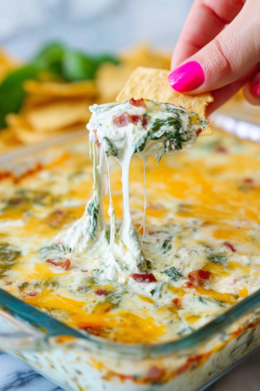 Bacon Spinach Artichoke Dip, creamy cheesy appetizer, easy bacon dip, hot spinach artichoke dip, crowd-pleaser appetizer - A white square glass dish shows a baked dip with a top layer of melted golden-yellow cheese, lightly browned in spots. Under the cheese, thick creamy white sauce mixed with green spinach leaves and small bits of reddish-brown bacon is visible. A woman's hand with pink painted nails holds a triangular beige chip pulling up a string of stretchy melted cheese from the dish, showing the layers clearly. The background is blurry with hints of green and more chips. The whole scene is set on a white marbled surface. photo taken with an iphone --ar 2:3 --v 7