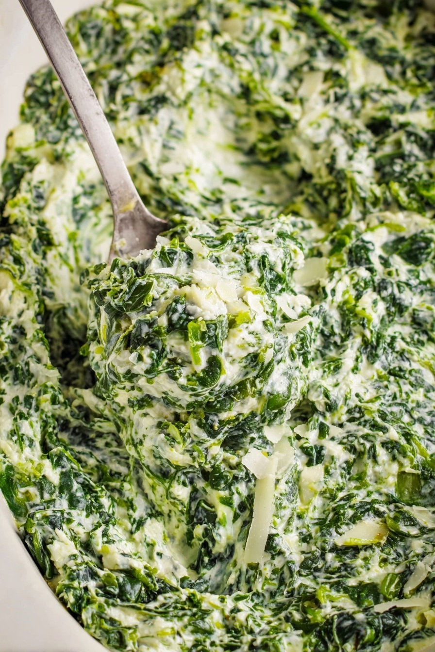 Creamed Spinach, Creamed Spinach Side Dish, How to Make Creamed Spinach, Easy Creamed Spinach Recipe, Creamed Spinach with Cheese - A close-up view of creamy spinach dip with visible layers of finely chopped green spinach mixed with white creamy sauce and small pieces of onion. The texture looks thick and rich with bits of grated white cheese sprinkled on top. A metal spoon scoops up a generous portion of the dip, showing the mix's dense, slightly chunky consistency. The dish sits on a white marbled surface that contrasts with the deep green and white colors of the dip. Photo taken with an iphone --ar 2:3 --v 7