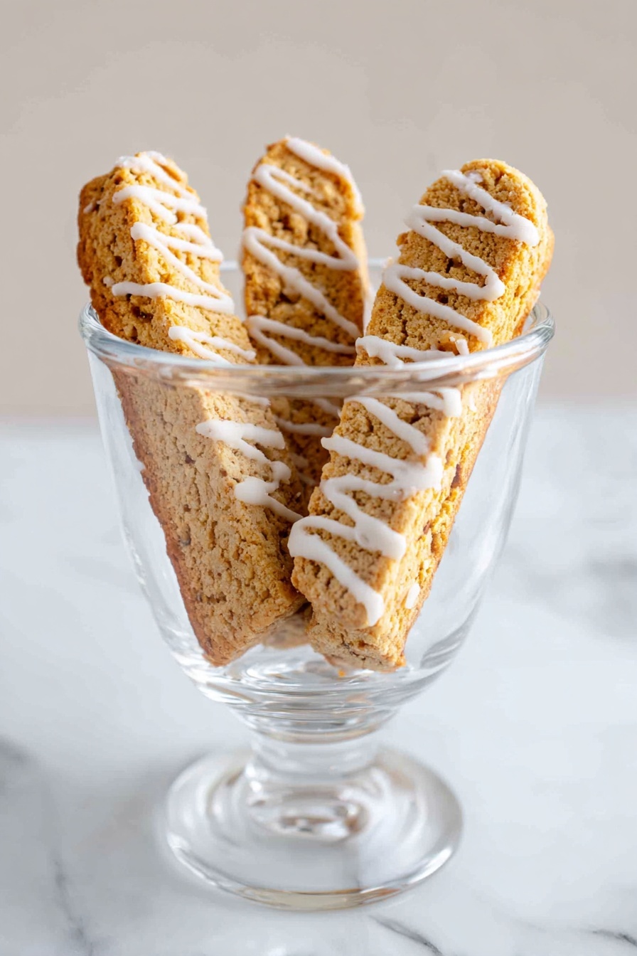 Cinnamon Vanilla Biscotti, biscotti recipe with cinnamon vanilla, how to make cinnamon vanilla biscotti, crunchy biscotti with vanilla and cinnamon, homemade cinnamon vanilla biscotti - The image shows a clear glass cup placed on a white marbled surface. Inside the cup, there are three crispy biscotti standing upright, each with a rough light brown texture. White icing is drizzled in thin stripes across each biscotti, adding contrast to their warm golden color. The background is plain and softly lit, emphasizing the cup and its contents. photo taken with an iphone --ar 2:3 --v 7