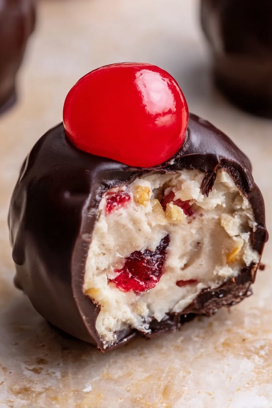 Cherry Bourbon Balls, Bourbon Truffles, Cherry Chocolate Bites, Holiday Treats, Boozy Candy - The image shows a close-up view of a small dessert ball with three layers. The outside layer is dark, smooth, and shiny chocolate coating covering the entire ball. Inside, the second layer is light-colored, creamy, and soft with some small red and golden brown bits mixed inside, giving it a textured look. On top of the chocolate layer, there is a bright red, glossy half of a cherry placed in the center. The ball sits on a light brown surface with a white marbled texture softly blurred in the background. photo taken with an iphone --ar 2:3 --v 7