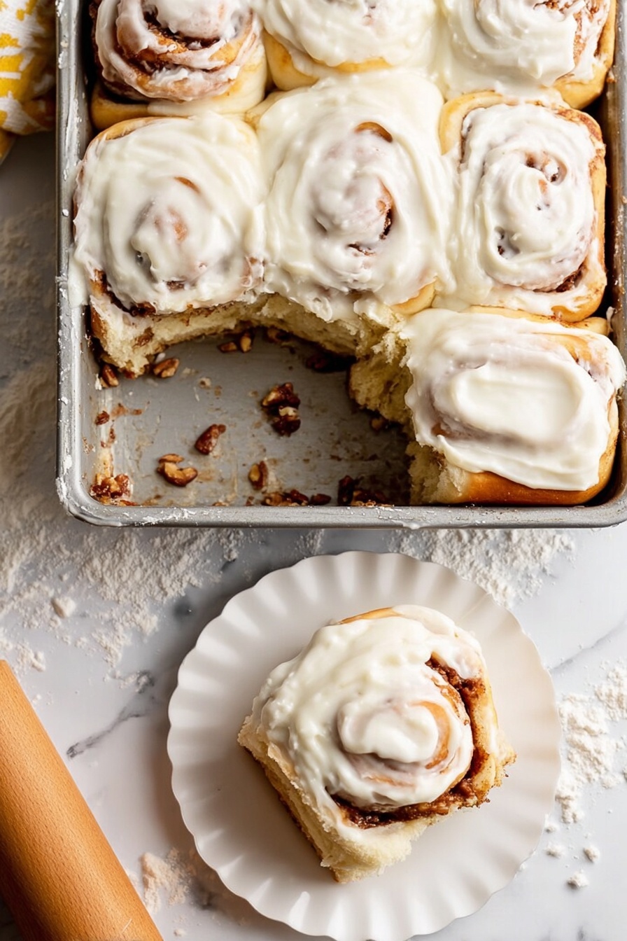 Soft Apple Cinnamon Rolls, apple cinnamon rolls, easy cinnamon roll recipe, homemade apple cinnamon buns, bakery-style cinnamon rolls - A metal baking dish holds six soft cinnamon rolls, with one removed showing the bottom layer coated in a sticky cinnamon filling and bits of nuts. Each roll has a creamy white icing thickly spread on top, swirled smoothly over the soft golden brown dough, with visible cinnamon spirals peeking through under the icing. One cinnamon roll sits on a small white scalloped plate in the lower right corner, showing its fluffy layered inside. The background is a white marbled surface lightly dusted with flour, with a wooden rolling pin nearby. photo taken with an iphone --ar 2:3 --v 7
