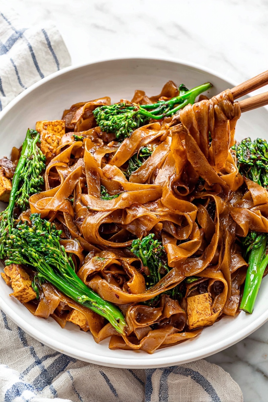Quick Thai Pad See Ew Stir Fry, Thai noodle stir-fry, easy Pad See Ew recipe, quick Thai dinner, authentic Thai stir-fry - A white bowl is filled with wide, flat noodles coated in a shiny dark brown sauce, layered thick, with some noodles lifted by wooden chopsticks to the right side of the image. Mixed in are pieces of light brown cooked tofu and bright green broccolini with textured small florets. The bowl sits on a white cloth with blue stripes on a white marbled surface. The colors show a contrast between the dark noodles, green vegetables, and pale tofu, creating a rich and fresh look. photo taken with an iphone --ar 2:3 --v 7