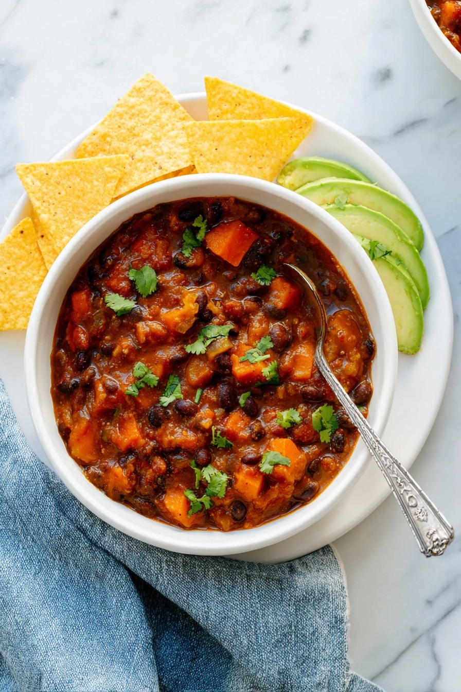 Vegetarian Chili, hearty vegetarian chili, easy vegetarian chili recipe, healthy chili recipe, flavorful vegetarian chili - A white bowl filled with thick chili containing black beans, orange carrot pieces, and chunks of sweet potato in a reddish-brown sauce, garnished with green cilantro leaves. On the right side inside the bowl are thin slices of light green avocado sprinkled with cilantro. The bowl sits on a white plate arranged with five triangular yellow tortilla chips around the left side. A silver spoon with a decorative handle rests inside the chili. The setting has a white marbled texture surface with a slightly crumpled blue cloth napkin next to the bowl. Photo taken with an iphone --ar 2:3 --v 7