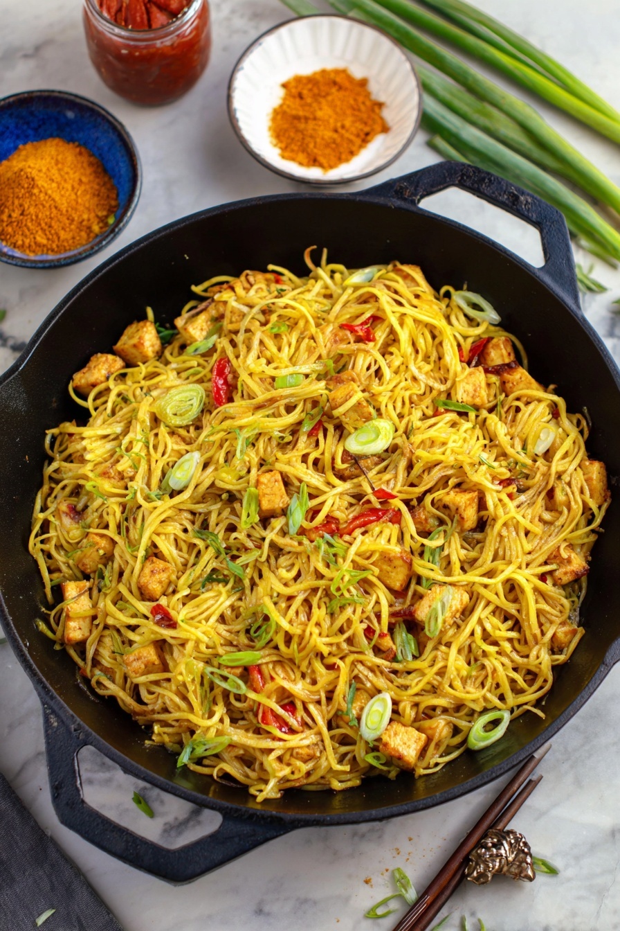 Quick and Easy Singapore Noodles, Singapore Noodles, Stir-Fry Noodles, Asian Noodle Recipes, Fast Weeknight Dinner - A black cast iron pan filled with several layers of yellow noodles mixed with small pieces of grilled tofu that show a light brown char, scattered with thin slices of green onions and small bits of red bell pepper. The noodles have a slight shine and look seasoned. Around the pan, there is a white bowl with orange powder, a jar with red sauce, a blue bowl with chopsticks placed on the side, and some green onion stalks on a white marbled surface. The scene has a mix of greens and yellows that make the dish look fresh and tasty photo taken with an iphone --ar 2:3 --v 7