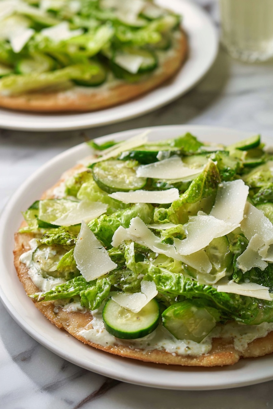 Viral Chicken Crust Caesar Salad Pizza, low carb chicken crust pizza, chicken salad pizza recipe, healthy chicken crust pizza, crispy chicken crust cauliflower pizza - The dish shows a flat round base with a light brown, slightly crispy texture. On top of the base, there is a layer of creamy white sauce spreading evenly. Above the sauce, there is a thick layer of green romaine lettuce pieces, mixed with round slices of light green cucumber scattered on top. Thin, irregular white shavings, likely cheese, are spread across the green vegetables. The dish is served on a simple white plate placed on a white marbled surface, with another similar dish visible blurred in the background. photo taken with an iphone --ar 2:3 --v 7